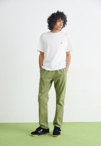 White cotton t-shirt with a small black logo, paired with light green pants and black sneakers with green accents, standing on a green surface.