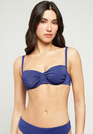 Frau mit langen dunklen Haaren, die einen strukturierten dunkelblauen Bikini-Oberteil und passende Unterteile vor einem einfarbigen hellen Hintergrund trägt.