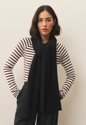 Ženska s kratkimi temnimi lasmi, oblečena v rjavo-belo črtasto majico z dolgimi rokavi, široke črne hlače in debel črni šal.