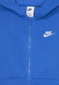 Kék cipzáros kapucnis pulcsi puha anyagból, bal mellen fehér Nike logóval, egyszerű dizájnnal, további minták nélkül.