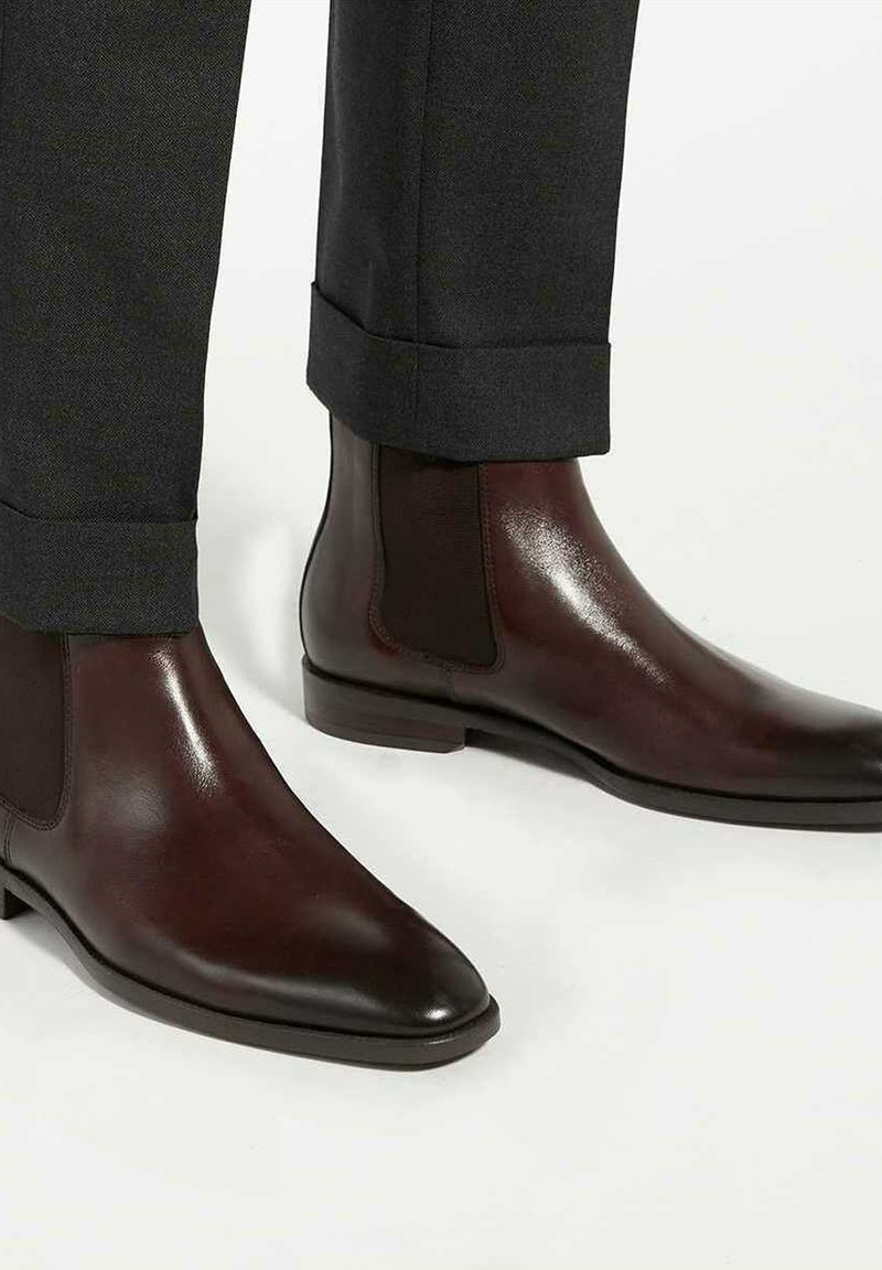 Bottes à cheville en cuir brun avec des panneaux latéraux élastiques, des embouts pointus et une finition lisse, assorties à un pantalon gris foncé bien taillé.