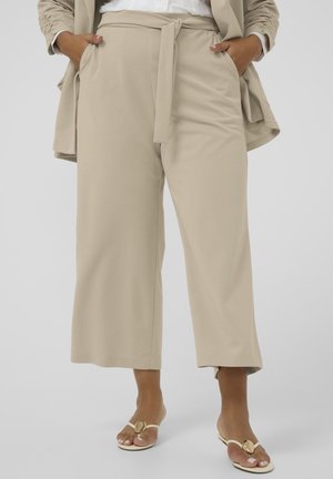Beżowe spodnie z wysokim stanem i szerokimi nogawkami z paskiem do wiązania, noszone z dopasowaną marynarką i białymi sandałami z odkrytymi palcami i złotymi akcentami.