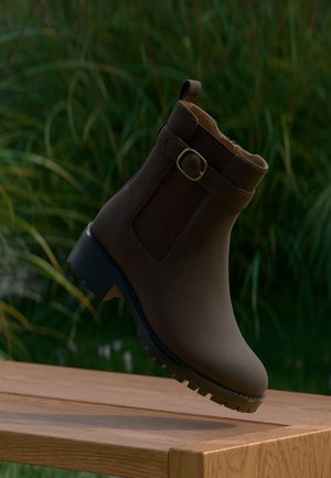 Botte marron à la cheville avec détail de boucle et semelle robuste flottant au-dessus d'une surface en bois avec des plantes vertes en arrière-plan.