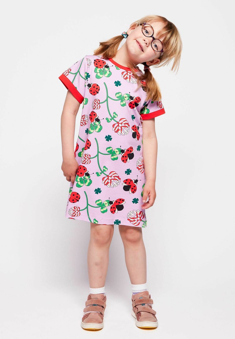Jeune fille portant des lunettes et une robe rose ornée de coccinelles, de fleurs et de trèfles, se tenant la tête inclinée et les mains détendues.