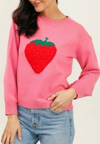 Pull en tricot rose avec un grand design de fraise rouge sur le devant. Col et poignets côtelés. Porté sur un jean bleu clair.
