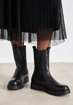 Czarne skórzane botki za kostkę z elastycznymi bocznymi panelami, masywnymi, bieżnikowanymi podeszwami oraz pętelkami do łatwiejszego zakładania, w zestawieniu z plisowaną czarną spódnicą.