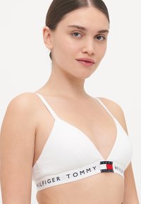 Bralette bianco a forma di triangolo realizzato in tessuto liscio, caratterizzato da un elastico con logo nei colori rosso, bianco e blu sul davanti.