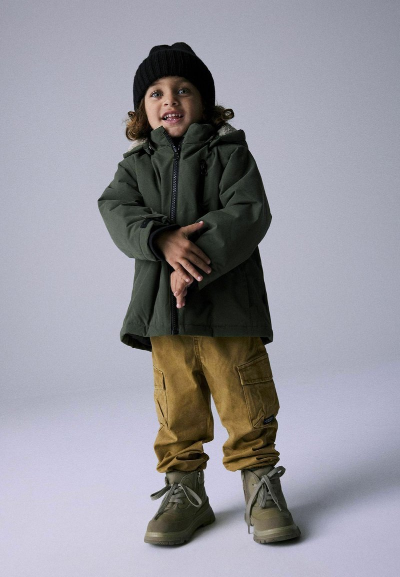 Giacca verde imbottita con zip, pantaloni cargo marroni con tasche e stivali grigi. Berretto di lana nera. Bambino in piedi con le braccia incrociate.