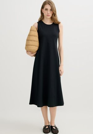 Mujer con vestido midi negro sin mangas sosteniendo un gran bolso beige tejido, de pie contra un fondo claro y liso.