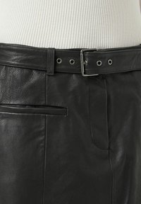 Sorte skinnbukser med teksturert finish, som har et belte med sølvfarge detaljer og en skrå frontlomme.