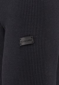 Schwarzer strukturierter Pullover mit einem gerippten Muster. Verfügt über ein rechteckiges schwarzes Etikett mit der Aufschrift "KEY LARGO", das am Stoff befestigt ist.