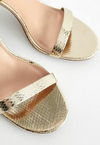 Guldmetallic hæle sandaler med et struktureret, slangeskinds mønster. Har en bred strop over tæerne og en glat, nude indersål.