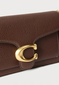 Καφέ πορτοφόλι από δερμάτινη υφή με καμπυλωτό χρυσό μεταλλικό κούμπωμα και εμφανείς μπεζ ραφές σε λευκό φόντο.