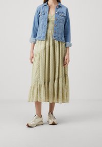 Giacca di jeans azzurro chiaro, abbottonata, con tasche; abito floreale verde a strati con orlo arricciato; scarpe sportive beige con suole strutturate.