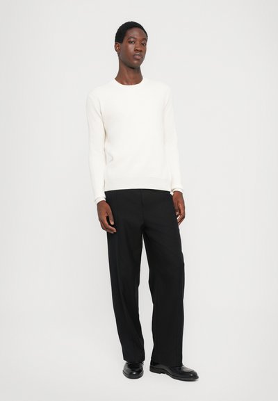 White ribbed sweater with long sleeves paired with black pleated trousers. Worn with black shoes. Simple, minimalistic design.
