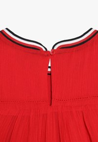 Vêtement rouge avec une texture côtelée, doté d'un col rond et d'une fermeture à boutons à l'arrière. Comprend une bordure rayée contrastante.