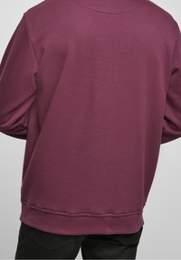 Persona vestida con un sudadera sencilla de manga larga de color púrpura oscuro y pantalones negros, vista desde atrás contra un fondo liso.