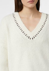 Biały sweter na drutach z głębokim dekoltem w kształcie litery V, z kontrastującą ciemną lamówką wzdłuż dekoltu. Tekstura jest miękka i gruba.