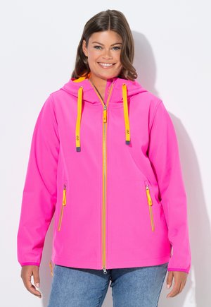 Femme souriante portant un sweat à capuche zippé rose vif avec des cordons et des détails de fermeture éclair jaunes, debout devant un fond blanc.