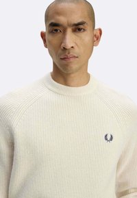 Maglione bianco sporco a coste con scollo rotondo e maniche raglan; presenta un logo blu navy sul petto. Texture morbida.