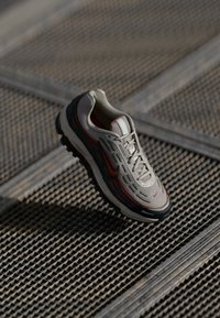 Nike sporta apavi ar sietu augšu gaiši pelēkā krāsā, akcentēti ar krāsainām līnijām oranžā un zaļā krāsā. Iekļauj teksturētu zoli un šņorēšanas sistēmu.