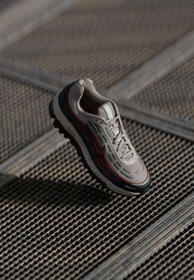 Nike sneaker, mille võrgusilma ülemine osa heledas hallis, millel on värvikas oranž ja roheline joonistus. Omab tekstureeritud talla ja paelade süsteemi.