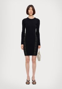 Black ribbed long-sleeve dress with gold eyelet detailing along the sides. Accompanied by a small silver mesh bag and black strappy sandals.