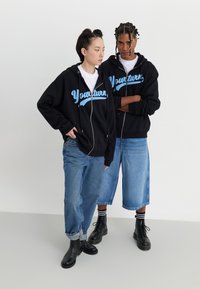Sorte hættetrøjer med blå skrift, der siger "Yourturn," båret over hvide skjorter. Modellerne sætter dem sammen med løse denimjeans og sorte støvler.
