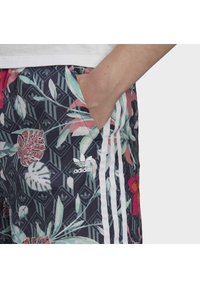 Handen i fickan, iklädd Adidas-shorts med blommönster och bladbilder samt vita tre ränder på mörk bakgrund.