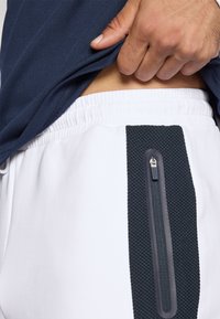 Białe dresy z szerokim, ciemnym, teksturowanym paskiem bocznym oraz kieszenią na zamek. W połączeniu z dopasowaną granatową bluzką, częściowo podniesioną.