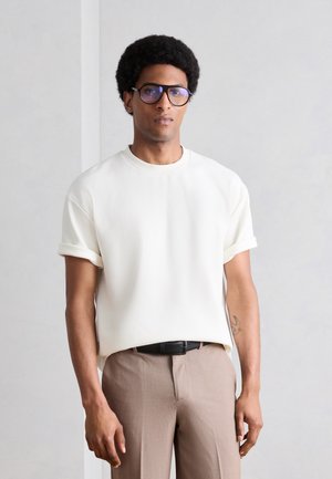 Νέος άνδρας με κόμμωση αφρο και γυαλιά φοράει λευκό πουκάμισο με κοντά μανίκια και μπεζ παντελόνι, στέκεται μπροστά σε έναν απλό ανοιχτό γκρι τοίχο.