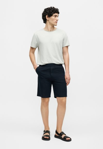 Νεαρός άνδρας με σγουρά μαλλιά που φοράει λευκό T-shirt, μαύρα σορτς και μαύρα σανδάλια, στέκεται με το ένα χέρι στην τσέπη και κοιτάζει προς τα δεξιά.