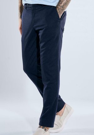 Man wearing navy blue trousers and white slip-on shoes, with tattooed arms and ankles visible, standing against a plain light background.