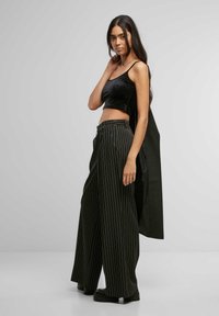 Černý sametový crop top, široké černé pruhované kalhoty a robustní černá obuv. Dlouhé vlasy, ukazující přiléhavou siluetu a uvolněný styl.