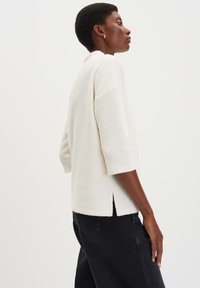 Donna che indossa un top bianco strutturato a tre quarti e pantaloni neri, in profilo contro uno sfondo chiaro e semplice.