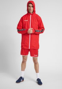 Rød vandtæt jakke med hætte, prydet med sorte chevronmønstre på ærmerne og en hvid lynlås, parret med matchende røde shorts.
