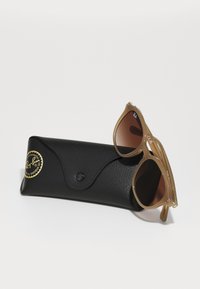 Ochelari de soare Ray-Ban cu ramele de culoare maro deschis și lentile degradate, expuși lângă un etui din piele neagră cu un logo aurit.