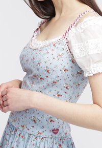 Donna che indossa un vestito floreale blu con spalline a quadretti rossi e maniche a sbuffo di pizzo bianco, con le mani unite in vita.