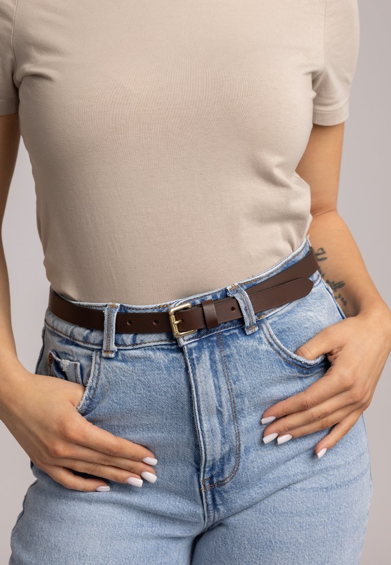 Personne portant une chemise beige clair à manches courtes rentrée dans un jean bleu clair avec une ceinture marron, les mains reposant dans les poches avant.