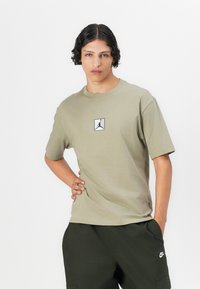 Oliven grøn bomulds t-shirt med korte ærmer, der har et centreret sort-hvidt Jordan-logo i en firkant. Modellen bærer mørkegrønne bukser.