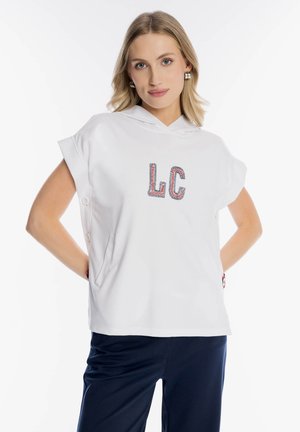 Blondynka w białej bezrękawniku z kapturem z napisem "LC" i granatowych spodniach, pozująca z rękami za plecami na białym tle.