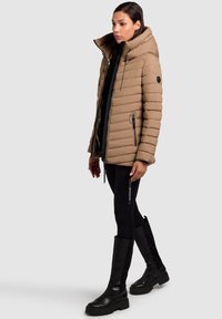 Beige Daunenjacke mit hohem Kragen, horizontaler Steppung, seitlichen Reißverschlusstaschen und einem Reißverschluss vorne, kombiniert mit schwarzen Leggings und knielangen Stiefeln.