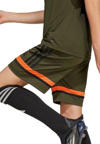 Olivgrüne Sportshorts mit schwarzen und orangefarbenen Streifen, kombiniert mit schwarzen knielangen Socken mit weißen Akzenten und einer enganliegenden Passform.