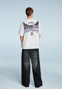 Bílé oversized tričko s černým a fialovým grafickým textem na zádech, kombinované s volně střiženými tmavými džínovými kalhotami s kapsami.