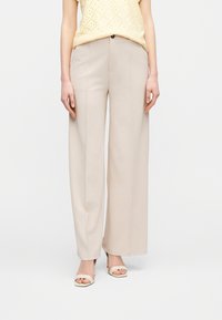 Γυναίκα που φοράει μπεζ ψηλόμεσο παντελόνι με φαρδιά μπατζάκια και μπλούζα χωρίς μανίκια σε κίτρινο χρώμα με τρύπες, συνδυασμένη με κρεμ ανοιχτά πέδιλα.