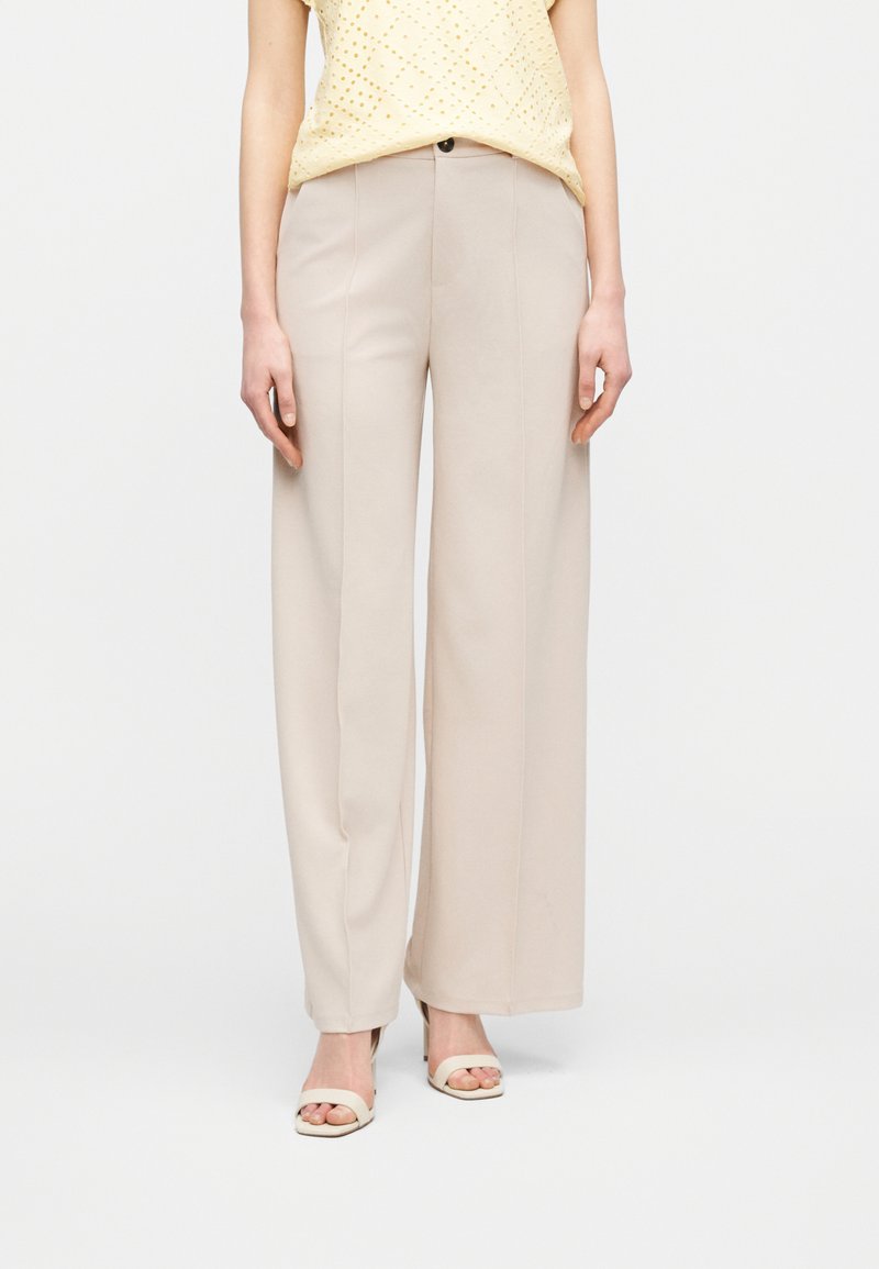 Γυναίκα που φοράει μπεζ ψηλόμεσο παντελόνι με φαρδιά μπατζάκια και μπλούζα χωρίς μανίκια σε κίτρινο χρώμα με τρύπες, συνδυασμένη με κρεμ ανοιχτά πέδιλα.