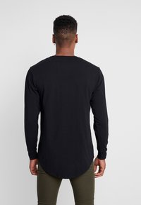 Černé tričko s dlouhými rukávy a kulatým výstřihem, měkká textura a zakřivený spodní okraj na zadní straně; spárované s olivově zelenými kalhotami.