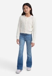 Cardigan bianco a maglia con motivo testurizzato e scollatura rotonda, abbinato a jeans svasati blu chiaro e scarpe da ginnastica bianche.