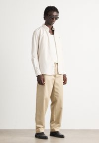 Camicia a righe beige con bottoni sopra una canotta bianca, abbinata a pantaloni larghi beige e scarpe nere, completata da occhiali da sole scuri.