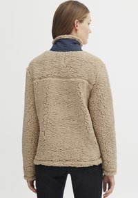 Veste en shearling beige avec une surface douce et texturée, dotée d'un col navy et d'une coupe décontractée et oversized. Un jean noir est porté en dessous.